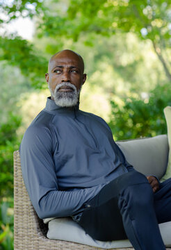 Mature man sitting on wicker patio wearing zipneck pullover and dark pants gazing at garden