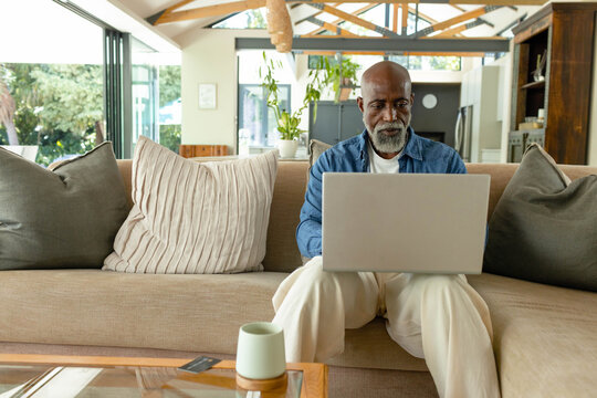 Senior African man sitting in living room on sofa holding laptop, focusing on screen near mug