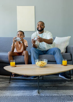 African American father and child sitting on gray sofa holding white controllers, snacking on chips