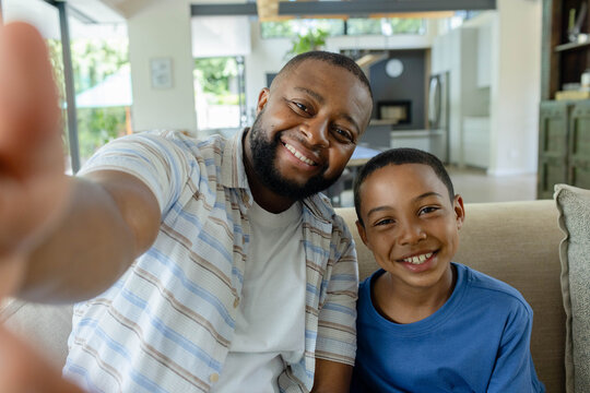 African American father and teen son sitting on beige couch at home reaching camera with smartphone