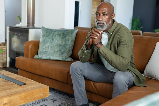 Mature African man sitting on brown leather sofa, clasping hands under chin in cozy living room