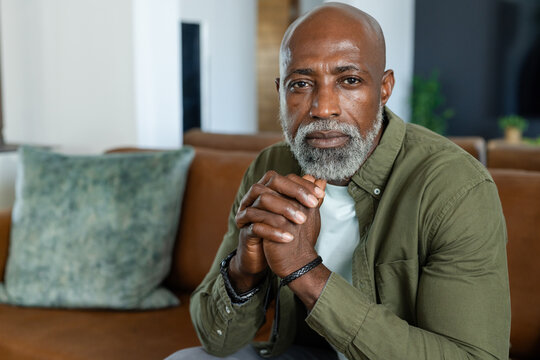 Senior African man sitting on sofa at home leaning clasping hands wearing olive shirt bracelet
