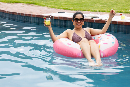 Woman reclining on pink donut float in backyard pool, wearing maroon bikini top, holding clear cup