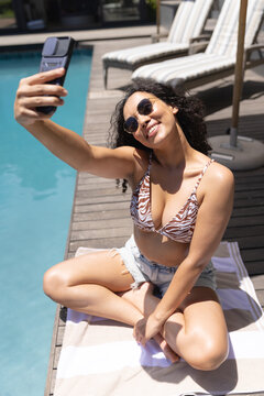 African American woman sitting in zebra bikini on wooden pool deck holding smartphone taking selfie