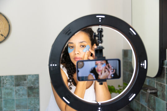 Adult African American woman fixing under-eye gel patches recording on phone by ring light at sink