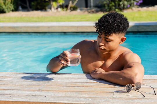 African American shirtless man leaning at backyard pool edge holding lemon glass, sunglasses nearby