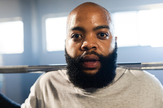 African American male lifting barbell across shoulders in gym weight room wearing grey t-shirt