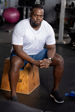 Male sitting on plyo box at gym wearing white tee, blue shorts, smartwatch, purple ball visible