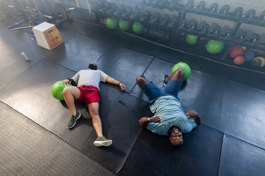 Two African men wearing workout clothes lying on weight room mats with green medicine balls