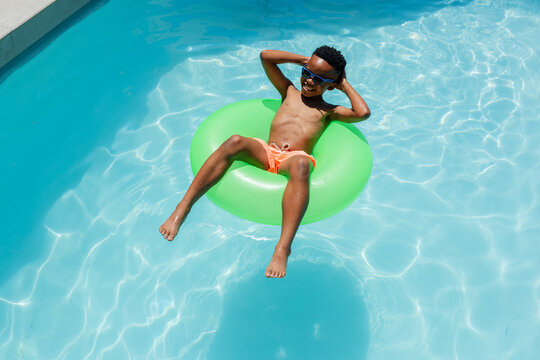 African youth male reclining on green inner tube in blue pool wearing orange trunks and shades