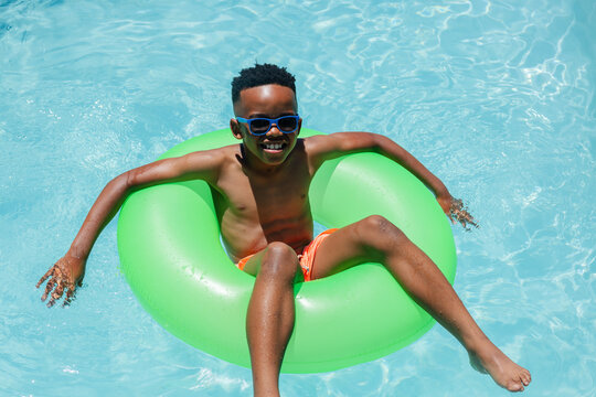 African American boy floating in green swim ring at pool, wearing blue sunglasses and orange trunks