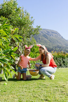 Woman and child girl wearing bunny ears collecting colorful eggs into wicker basket on lawn