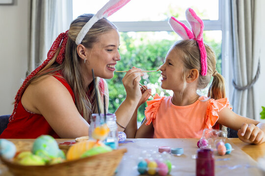 Adult woman and girl wearing bunny ears holding brush painting eggs and face at wooden table