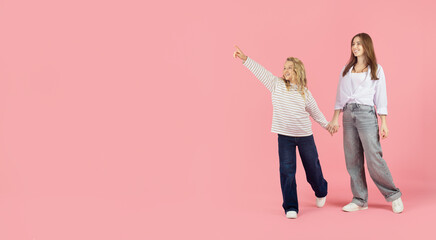 Fototapeta premium Little girl pointing forward on pink studio background and holding mothers hand, expressing curiosity and positive family lifestyle moment, copy space