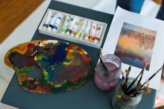 Artist palette is resting on gray-blue table with paint tubes, jars of brushes and reference print