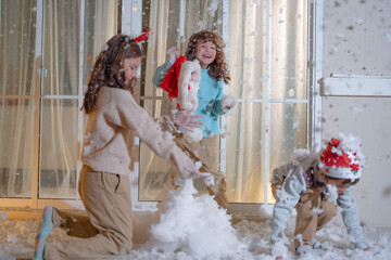 Three kids dressed in festive Christmas clothing enjoy playing with artificial snow on a cozy balcony, capturing the warmth and fun of the holiday season at home in a joyful moment. © ultramansk