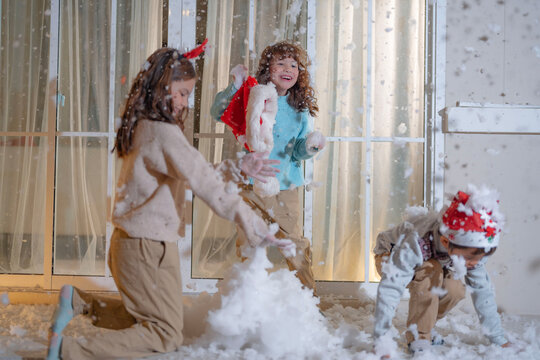 Three kids dressed in festive Christmas clothing enjoy playing with artificial snow on a cozy balcony, capturing the warmth and fun of the holiday season at home in a joyful moment.