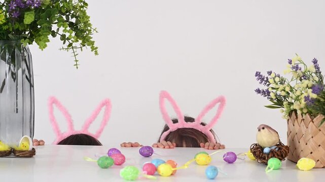 A boy and girl wearing bunny ears and an Easter bunny costume play hide and seek while peeking out from behind a table decorated with flowers and colorful Easter eggs.