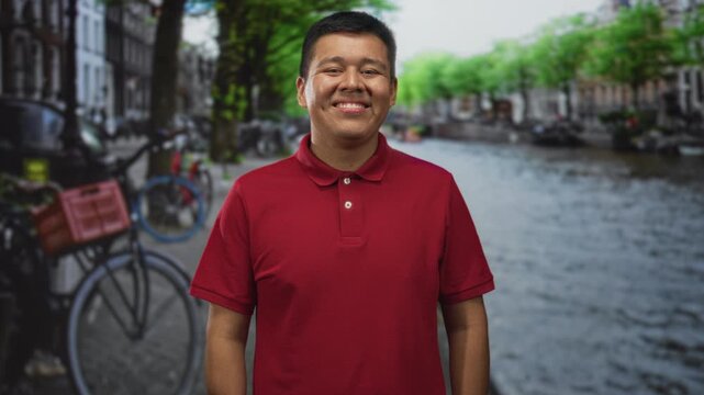 Man smiling with visible face, wearing a red polo while standing beside an amsterdam canal with parked bicycles and waterway; happiness travel.