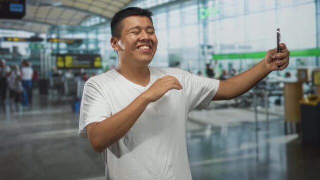 Hispanic man smiling with eyes closed arm extended holding smartphone taking a selfie in busy airport terminal wearing white t shirt; travel joy spontaneity.