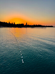 Fototapeta premium Early sunrise at Issyk Kul Lake in Kyrgyzstan. Warm golden sky reflecting in calm water with silhouettes of trees and pier on horizon.