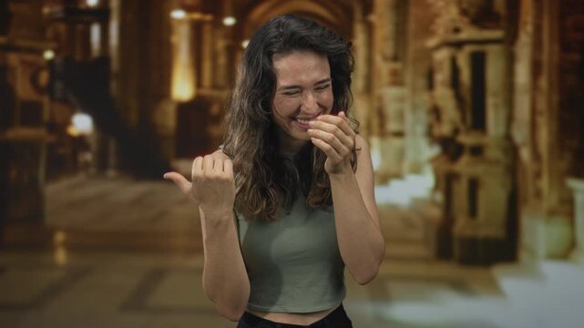 Woman covers mouth with hand and points thumb behind her in church building while wearing green top; amusement.