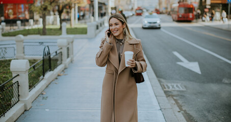 Obraz premium Woman talks on phone while walking on city street with coffee in hand during day
