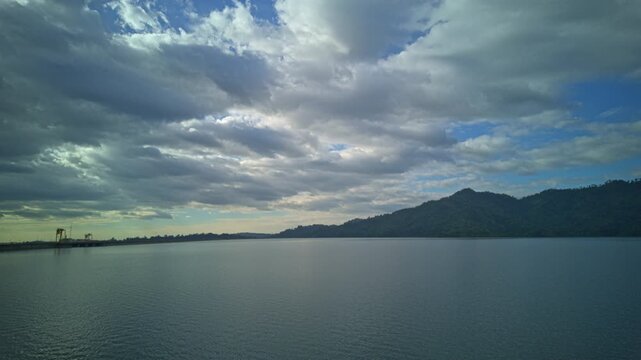 Time lapse stunning landscape view of Khun Dan Prakan Chon Dam showcasing calm blue waters reflecting dramatic white clouds and lush green mountains, creating a peaceful natural panorama.