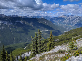 Obraz premium Wide view of rugged mountains and a deep alpine valley with dense evergreen forest, rocky foreground & dramatic cloud shadows. Serene, expansive landscape ideal for hiking, travel & outdoor adventure.