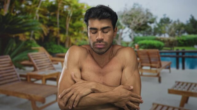 Man bare chest wraps arms around torso beside sunlit deckchair against building wall; cold discomfort.