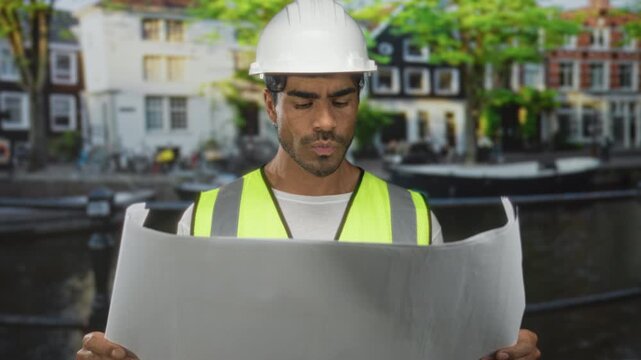 Young man in white hardhat and reflective vest reads large blueprint on street near canal by building facade; focus.
