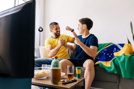 Brazilian father and teenage son watching football match together at home celebrating exciting moments. Family bonding through sports entertainment sharing emotion, support and generational connection