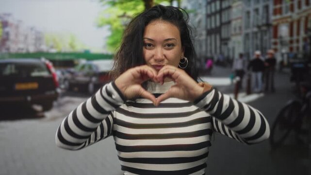 Woman wearing striped turtleneck makes heart gesture with hands in a busy city street, smiling and holding heart near face; joy love affection.