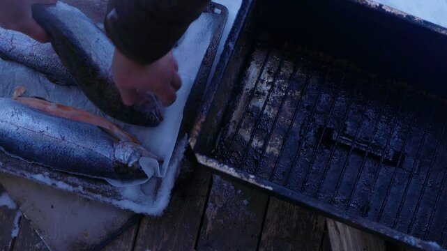 Fresh whole fish placed on grill rack inside metal smoker outdoors in winter. Preparing for hot smoking process in snowy conditions. Traditional outdoor cooking and fish preservation concept