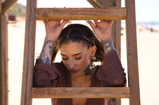A beautiful young woman in a brown dress rests her arms on the steps of the wooden ladder of the lifeguard hut on the beach. The woman is sad and depressed and is going through a difficult time.