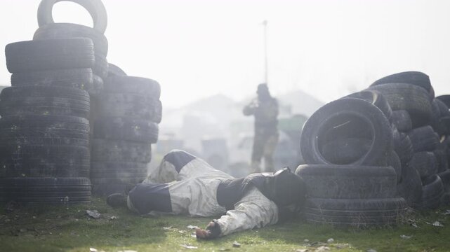 A player lies on the ground, eliminated during a paintball or air soft game on a tactical field. Another participant stands ready in the hazy background, continuing the intense combat simulation.