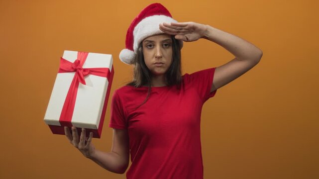 Woman holding white gift box with red ribbon and saluting in orange studio; festive cheer holiday giving.