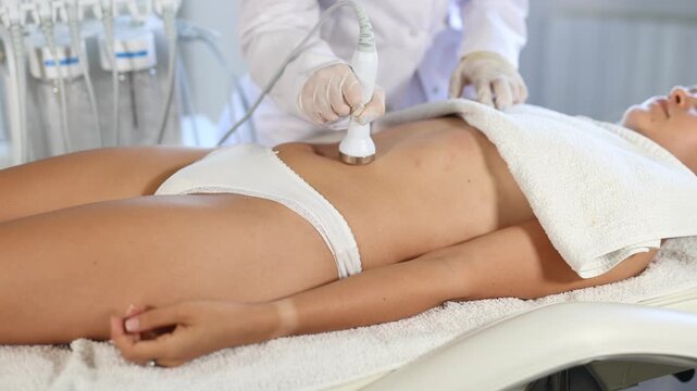 Female patient undergoing ultrasonic cavitation procedure on belly at modern aesthetic medicine clinic to reduce cellulite and adipose fat, improve body shape and reduce circumference. High quality 4k