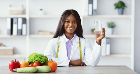 Naklejka premium A woman in a lab coat holds a glass of water while sitting at a table with various fresh vegetables and fruits. She is promoting a healthy lifestyle in a bright office setting.
