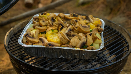 Grilled vegetables and mushrooms in aluminum tray on barbecue grill outdoors surrounded by natural forest setting