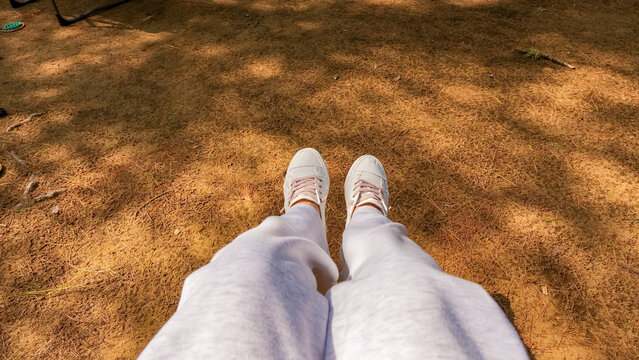 Person wearing white sneakers and gray sweatpants sitting on brown ground with scattered leaves and dappled sunlight filtering through trees