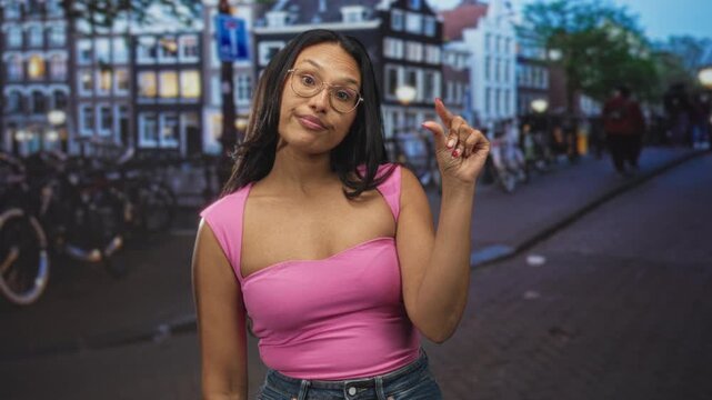 Woman in pink top holds thumb and forefinger close in a small pinch gesture on a cobbled street with bicycles and canal buildings; playful attitude.