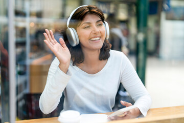 Smiling person wearing headphones and waving while sitting at a table in a busy cafe © Iryna