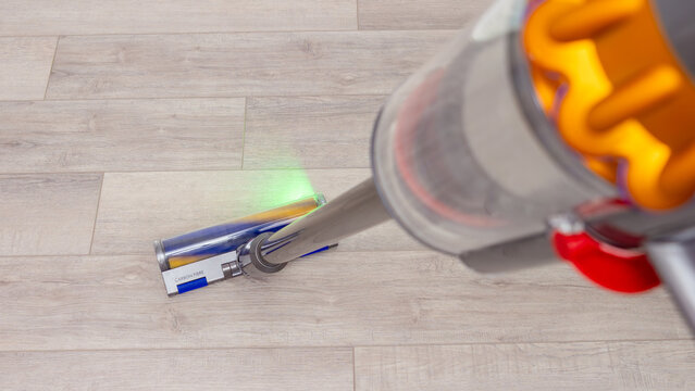 Cordless stick Dyson vacuum cleaner cleaning hardwood floor in home interior with green light detail, everyday housekeeping, organized living, hygiene and comfort.
