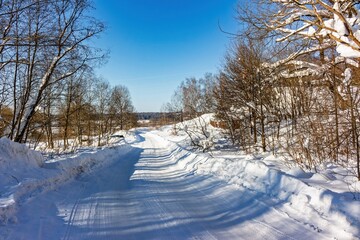 Obraz premium Serene winter wonderland with a pristine snow-covered road winding through bare trees under a brilliant blue sky. Long shadows create depth on a sunny day