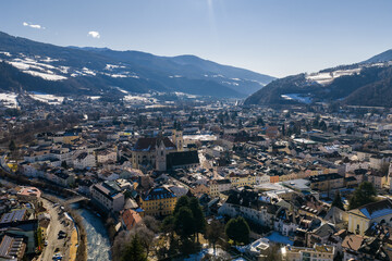 Aerial view of Brixen in South Tyrol, Italy, with Brixen Cathedral, pastel buildings, winding streets, Eisack River, small bridges, and snow dusted Dolomite foothills.