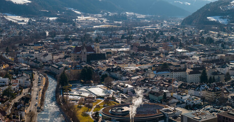 Obraz premium Aerial view of Brixen, Italy, shows the Eisack River, Brixen Cathedral, terracotta roofs, solar panels, and snow dusted Alpine foothills on a calm late winter day.