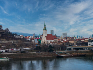 Fototapeta premium St. Martin's Cathedral with green and gold spire rises over Bratislava. Danube River, bridges, embankments, modern high rises, and winter bare trees appear in cool daylight.