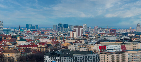 Obraz premium Aerial view of Bratislava, Slovakia, shows historic rooftops, the green capped Old Town Hall tower, church spires, and modern glass high rises under a bright partly cloudy sky.