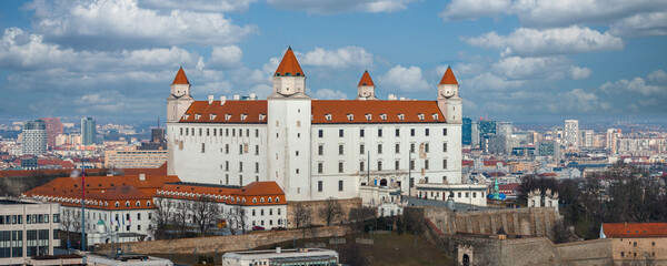 Bratislava Castle with four corner towers and terracotta roof stands on a hill above Bratislava, with old walls, palace buildings, and modern skyline in clear day © Aerial Film Studio